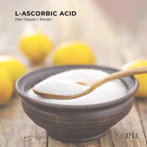 Bowl filled with white crystalline Vitamin C powder with wooden spoon resting on top, surrounded by fresh yellow lemons in soft focus, promotional text highlighting L-Ascorbic Acid purity and orku branding