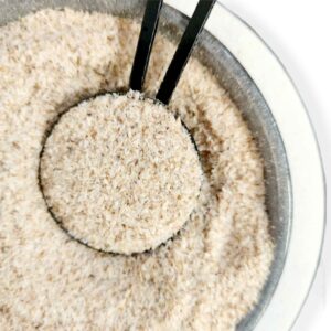 Close-up view of bowl filled with finely ground beige psyllium husk, black measuring spoon scooping portion from center, showcasing light flaky texture and serving method for digestive health supplement