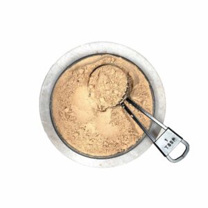 Ceramic bowl filled with finely ground light brown maca powder, metal measuring spoon labeled 1 TBSP resting on top, showcasing smooth texture and natural presentation of this energizing root supplement