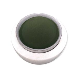 Small round dish filled with finely ground dark green spirulina powder, placed on a white ceramic base, showcasing smooth texture and vibrant color typical of nutrient-rich algae supplements
