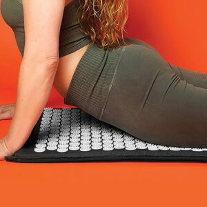person in greenish-brown athletic outfit lying face down on acupressure mat with white spiked discs, placed on bright orange surface for muscle stimulation and wellness