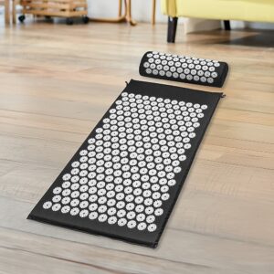 Black acupressure mat and pillow set with white spike discs laid out on wooden floor in living room; yellow couch and furniture in background