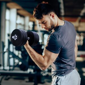 Person performing a bicep curl with a dumbbell in a gym environment wearing a dark t-shirt and light pants with fitness equipment in the background
