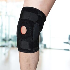 Leg wearing black knee brace with red patella ring and Velcro straps; gym background suggests rehab or training use