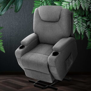 Grey upholstered massage recliner chair with a cushioned headrest, padded armrests, and built-in cup holders on both sides, placed on a dark wooden floor against a textured wall with green leafy plants in the background. The chair sits on a black metal base and is designed for comfort and relaxation.
