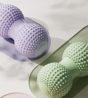Two peanut-shaped massage rollers with dual spiky ball ends connected by a central grip, shown in pastel purple and green. The textured nodules are designed for deep tissue massage and trigger point therapy, placed on flat surfaces with soft shadows against a minimalist background.