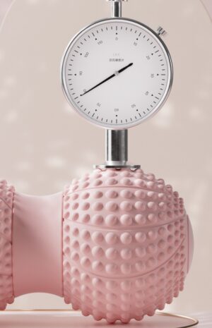 Close-up of a pink peanut-shaped massage ball roller with raised spiky texture being tested under a metal pressure gauge. The gauge is mounted vertically and presses down on the rounded end of the roller, demonstrating strength and compression resistance. The surface shows evenly spaced nodules designed for deep tissue massage, and the soft neutral background highlights the product’s durability and high-pressure tolerance.