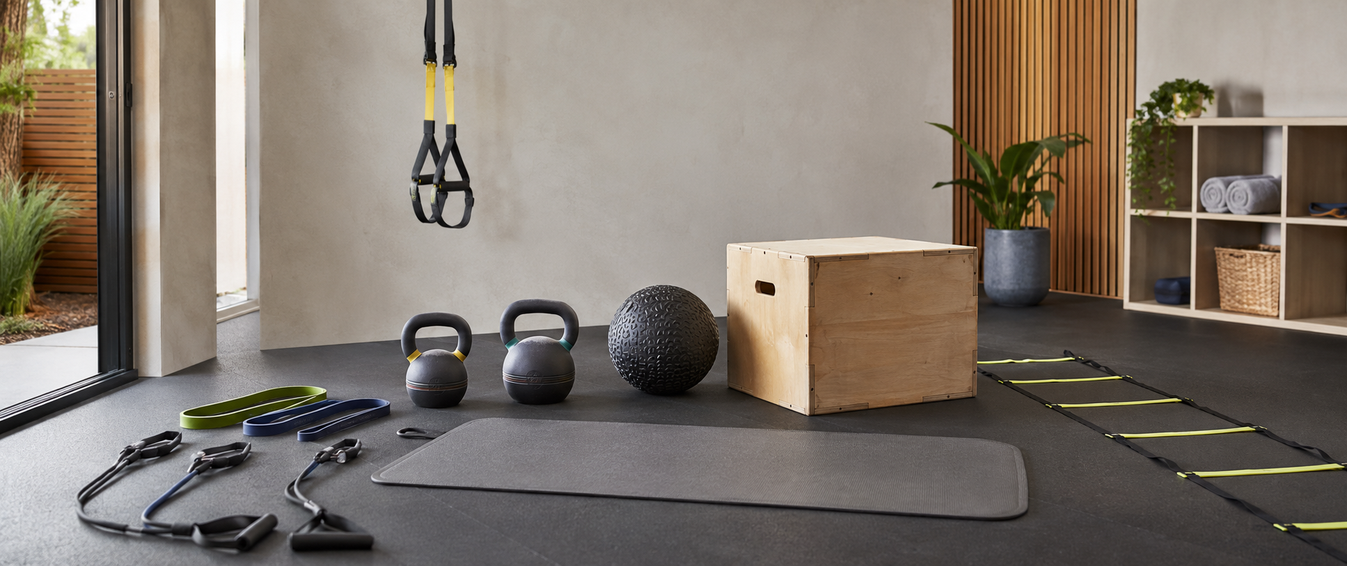 Functional training equipment in a home gym with kettlebells, resistance bands, suspension straps, a medicine ball, plyometric box and agility ladder